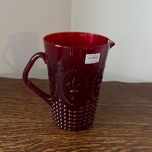 May include: A red glass pitcher with a textured design and a handle. The pitcher has a label that reads "IVIMA MADE IN PORTUGAL".