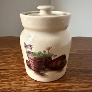 May include: A cream-colored ceramic jar with a lid. The jar features a painted scene of a bird, flowers, and potted plants. The jar is sitting on a wooden surface.