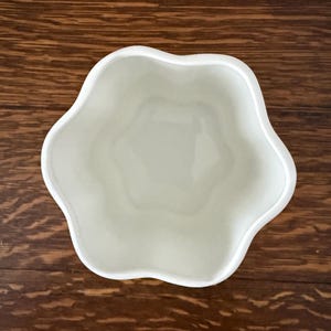 May include: A white ceramic bowl with a distinct five-petaled flower shape viewed from directly above. The interior of the bowl features smooth, curved walls that follow the scalloped rim, creating a symmetrical and open vessel design.