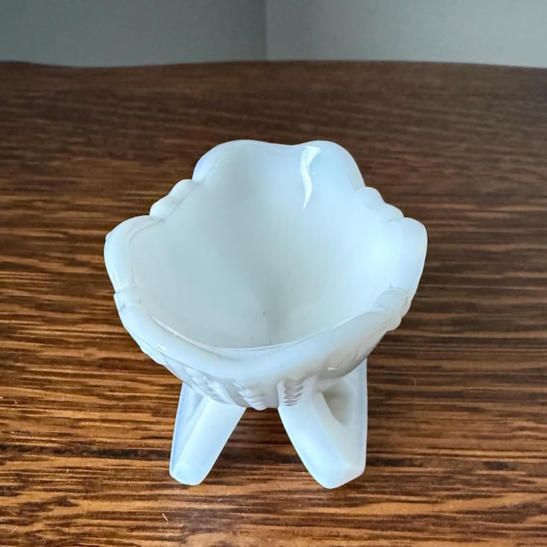 May include: A small, white, glass dish with a scalloped rim and three legs. The dish has a smooth, opaque appearance and is likely vintage. The dish is sitting on a wooden surface.