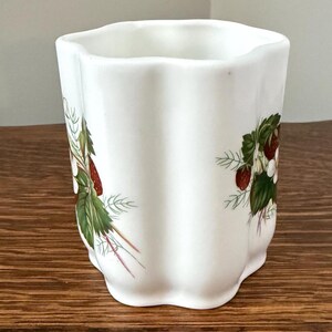May include: A white ceramic tumbler with a scalloped, wavy rim and vertical fluted sides. The exterior features a botanical illustration of red strawberries, green leaves, and small white flowers arranged in clusters around the body.