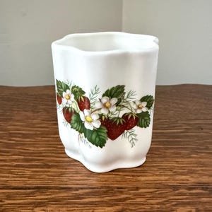 May include: A white ceramic tumbler with a scalloped rim and base, featuring a botanical illustration of red strawberries, green leaves, and white flowers with yellow centers wrapped around the exterior of the cylindrical vessel.
