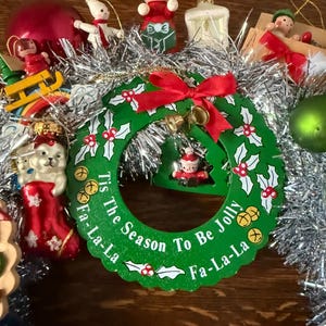 May include: A festive Christmas wreath with a green base, adorned with holly leaves, red berries, and a red bow. The wreath features the text "Tis The Season To Be Jolly Fa-La-La". Various ornaments surround the wreath.