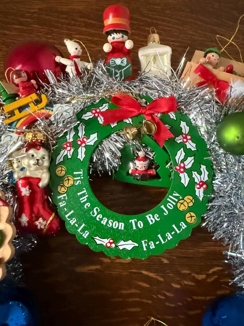 May include: A festive Christmas wreath with a green base, adorned with holly leaves, red berries, and a red bow. The wreath features the text "Tis The Season To Be Jolly Fa-La-La". Various ornaments surround the wreath.