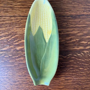 May include: A green and yellow ceramic corn on the cob serving dish. The dish is shaped like a corn cob and has a green husk and yellow kernels.