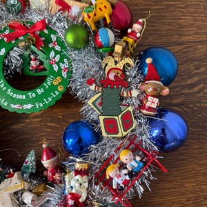 May include: A festive Christmas wreath with silver tinsel and various ornaments. The wreath includes miniature wooden toys, red and blue baubles, and a green wreath with the text "The Season To Be Jolly".