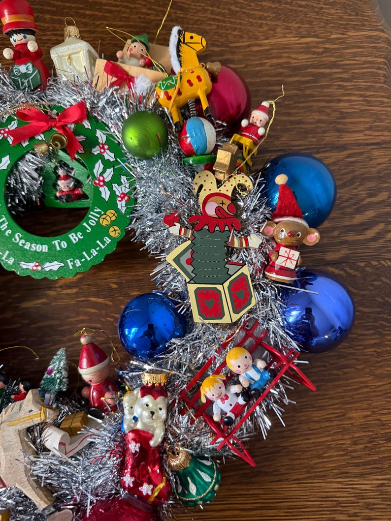 May include: A festive Christmas wreath with silver tinsel and various ornaments. The wreath includes miniature wooden toys, red and blue baubles, and a green wreath with the text "The Season To Be Jolly".