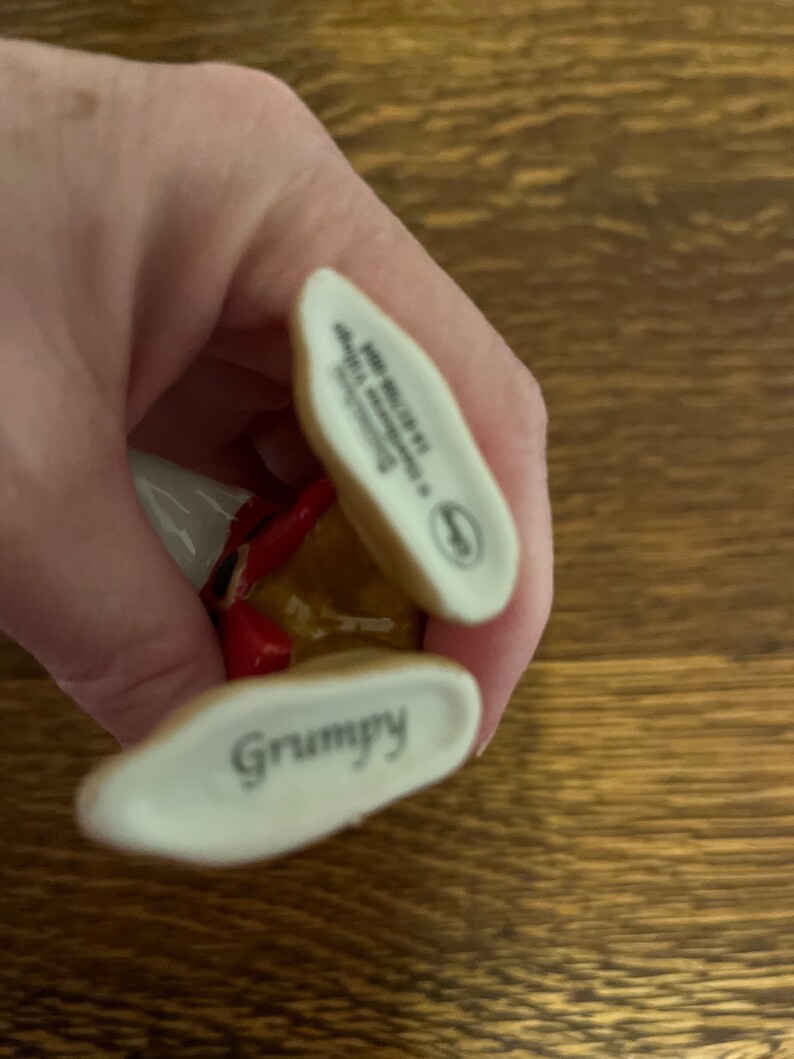 Może przedstawiać: Ceramiczna figurka Grumpy'ego z filmu Disneya Śnieżka i siedmiu krasnoludk&oacute;w. Figurka jest pomalowana na brązowo, czerwono i biało. Figurka ma na sobie czerwony kapelusz i brązowy płaszcz. Figurka stoi na białej podstawie z napisem "Grumpy". Figurka jest oznaczona na spodzie napisami "Jim Shore" i "Enesco".