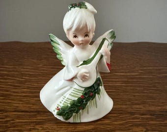 May include: A ceramic angel figurine in a white dress with green and red accents, holding a musical instrument. The angel has white wings and hair, and is decorated with a holly garland. A festive holiday decoration.