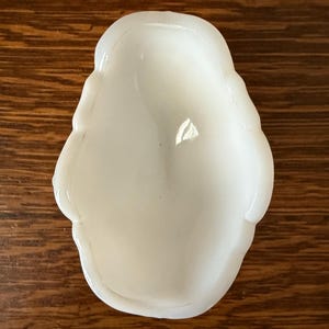 May include: A small, white, opalescent dish with a rounded, irregular shape. The dish has a smooth, glossy surface and a shallow interior. The edges are slightly raised and have a wavy design. The dish is sitting on a wooden surface.