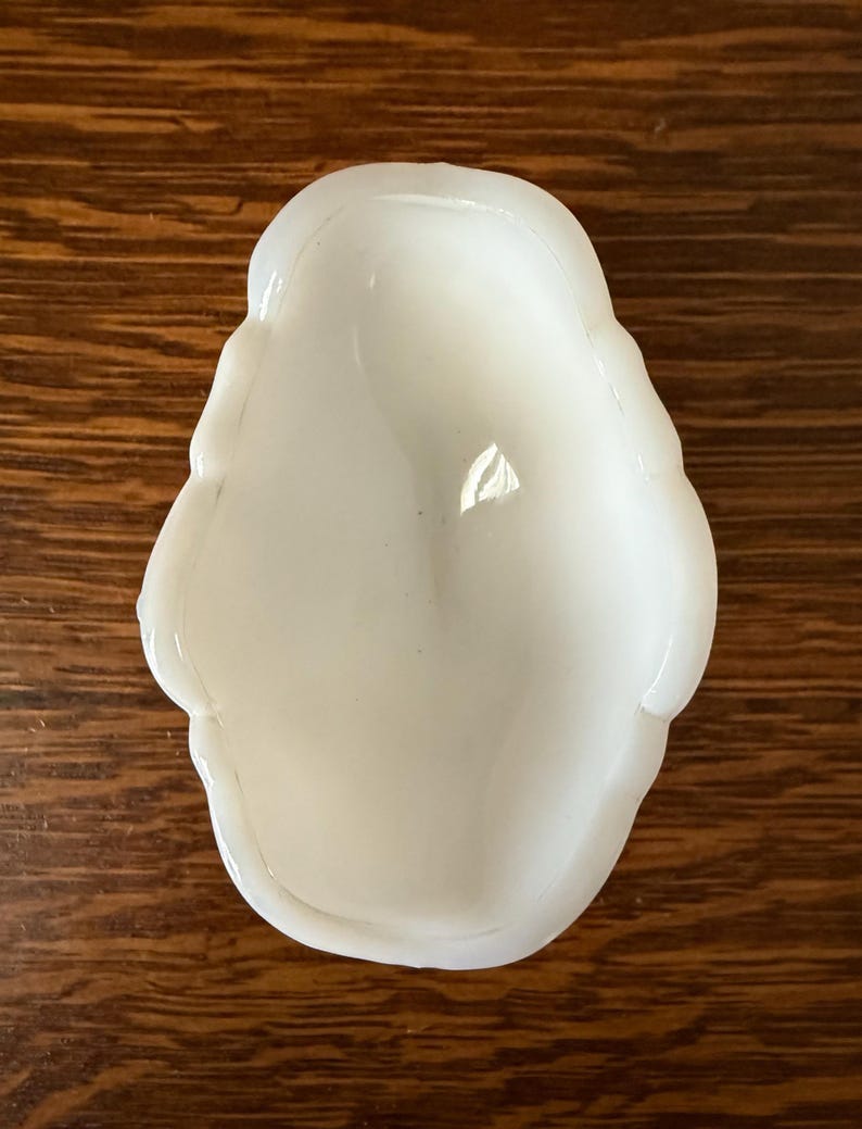 May include: A small, white, opalescent dish with a rounded, irregular shape. The dish has a smooth, glossy surface and a shallow interior. The edges are slightly raised and have a wavy design. The dish is sitting on a wooden surface.