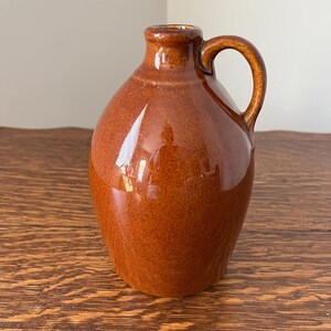 May include: A small, brown ceramic jug with a handle. The jug has a rounded body and a narrow neck. The surface is glossy and reflects light. The jug is likely used for storing liquids and has a rustic, vintage aesthetic.