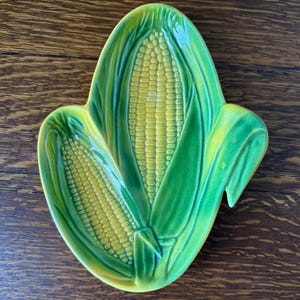 May include: A green and yellow ceramic dish shaped like two ears of corn. The dish is perfect for serving corn on the cob.