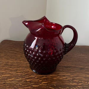May include: A red glass pitcher with a textured, bumpy surface. The pitcher has a wide mouth and a handle.