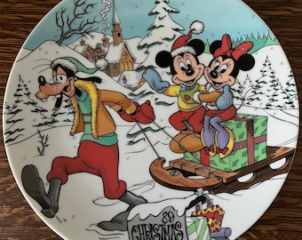 May include: A white ceramic Christmas plate featuring Mickey Mouse, Minnie Mouse, and Goofy. Mickey and Minnie are riding a sled pulled by Goofy, who is wearing a red and green winter outfit. The plate has a snowy winter scene with a village in the background and the text '89 Christmas' on a sign.