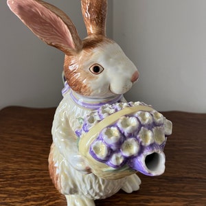 May include: A ceramic bunny teapot with brown and white fur, holding a purple and yellow flower basket. The spout is shaped like a flower.