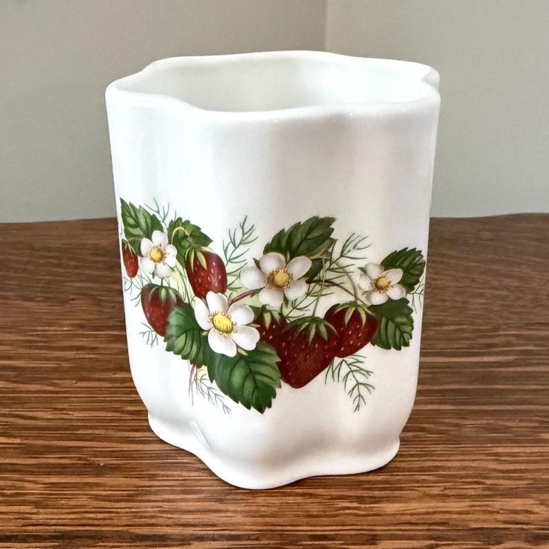 May include: A white ceramic utensil holder with a scalloped rim and a wavy body. The exterior features a botanical illustration of red strawberries, green leaves, and white strawberry blossoms arranged in a horizontal band around the center.