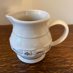 May include: Cream-coloured ceramic jug with a curved handle and spout. The jug features a basket weave design with green floral accents around the base and on the body. A classic piece for serving beverages.