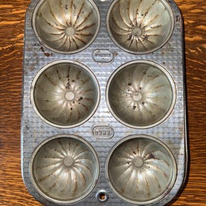 May include: A vintage silver-colored metal baking pan with six individual molds. The molds have a fluted design and are slightly rounded. The pan has the text "EKCO" and "MADE IN USA" printed on it.