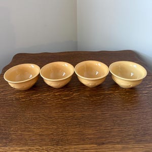 Peut inclure: Quatre petits bols ronds orange clair avec un motif texturé en forme de panier sur les côtés. Les bols sont disposés en rangée sur une surface en bois foncé, avec un fond neutre.