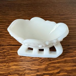 May include: A white, decorative dish with a scalloped rim and a pedestal base. The dish has a cloud-like shape and is made of a glossy, opaque material. The base has a rectangular design with three openings.