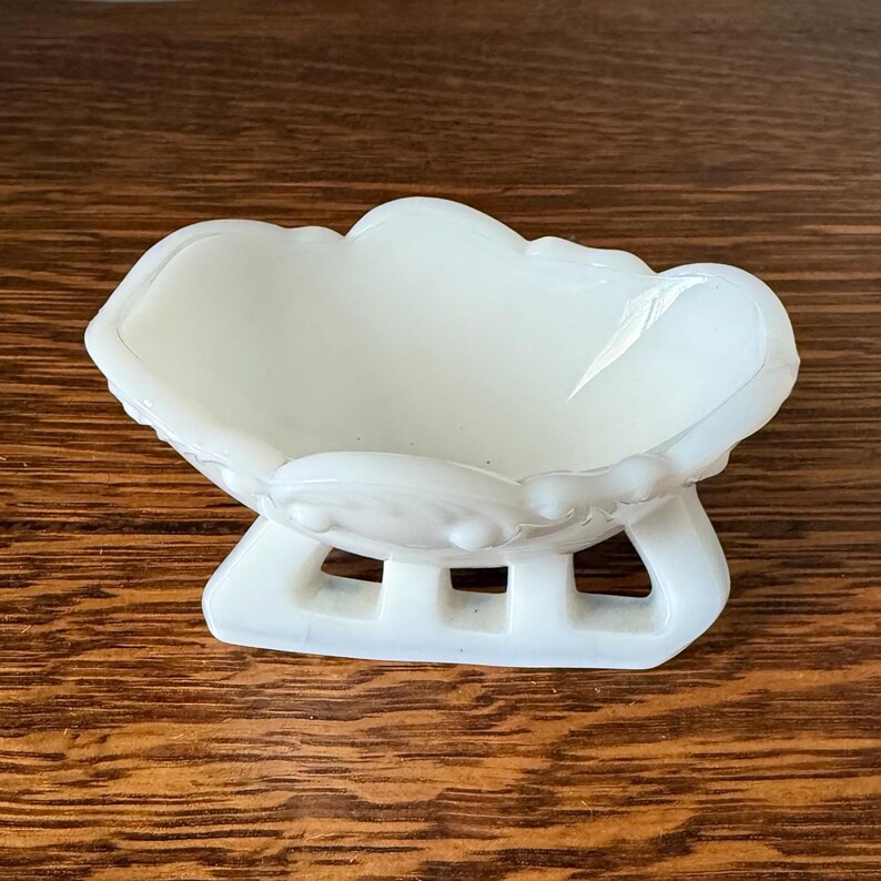 May include: A white, decorative dish with a scalloped rim and a pedestal base. The dish has a cloud-like shape and is made of a glossy, opaque material. The base has a rectangular design with three openings.