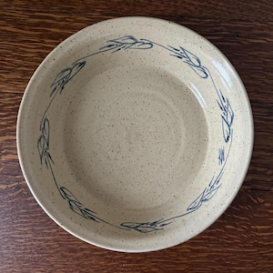 May include: A speckled, beige ceramic plate with a blue wheat stalk design around the rim. The plate has a wide, shallow shape, suitable for serving. The plate is on a wooden surface.