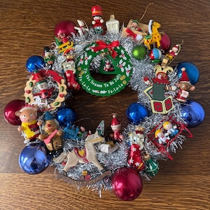 Handmade 12 inch vintage Christmas ornament wreath featuring assorted small figurines, glass ornaments, and silver tinsel garland arranged in a circular wreath form.