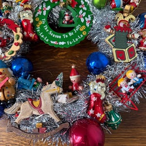 May include: A festive Christmas wreath with a silver tinsel base, adorned with various ornaments. The wreath features a green circular centerpiece with the text "Tis The Season To Be Jolly Fa-La-La". Ornaments include a rocking horse, red and blue baubles, and wooden figurines.