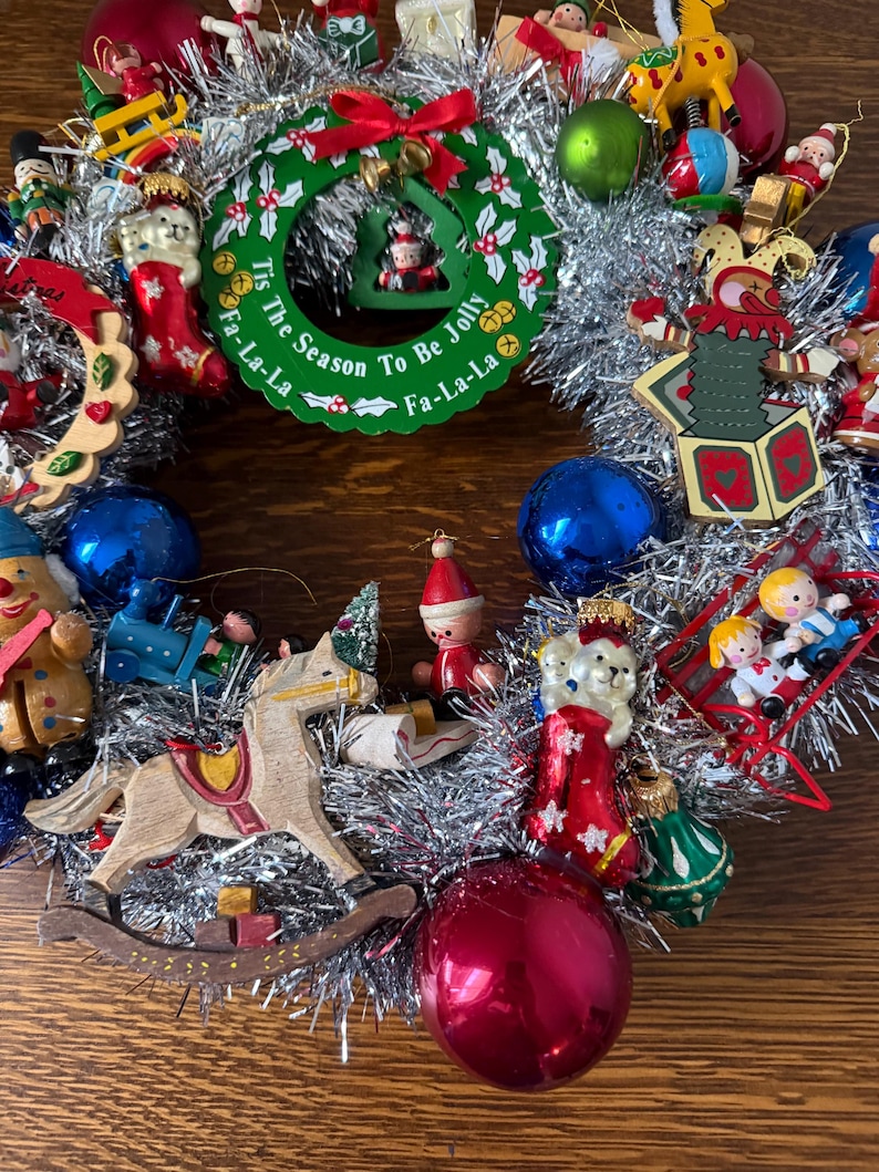 May include: A festive Christmas wreath with a silver tinsel base, adorned with various ornaments. The wreath features a green circular centerpiece with the text "Tis The Season To Be Jolly Fa-La-La". Ornaments include a rocking horse, red and blue baubles, and wooden figurines.