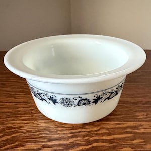 May include: White ceramic bowl with a wide rim and a decorative band of blue floral and leaf patterns around the exterior. The bowl has a smooth, glossy finish and is set on a wooden surface.