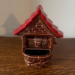 May include: A ceramic wishing well planter with a red roof and brown brick-like detailing. The well features a bucket and rope, with a dark opening for planting. The planter is designed to sit on a flat surface.