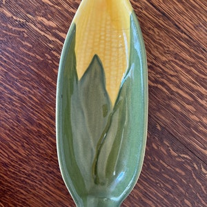 May include: A green and yellow ceramic dish shaped like an ear of corn. The dish is glazed and has a glossy finish.