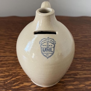 May include: A tan ceramic coin bank with a handle and a slot for inserting coins. The bank has a rounded body and a blue emblem that reads "UHL Collectors Inc." and "Huntingburg, Indiana."