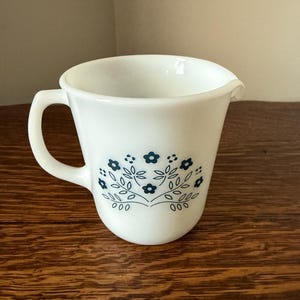 May include: White ceramic creamer with a handle and a spout. The creamer features a blue floral design on the front. The handle is curved, and the spout is slightly angled. The creamer is likely vintage and suitable for serving cream or milk.