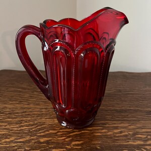 May include: A red glass pitcher with a scalloped rim and a textured design. The pitcher has a handle and a rounded base.