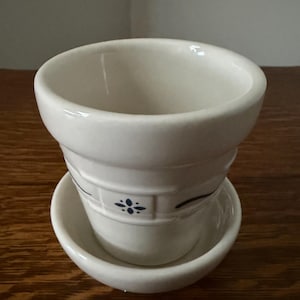 May include: A white ceramic planter with a matching saucer. The planter has a slightly flared shape with a decorative band and a blue floral design. The saucer is round and shallow. The items are on a wooden surface.
