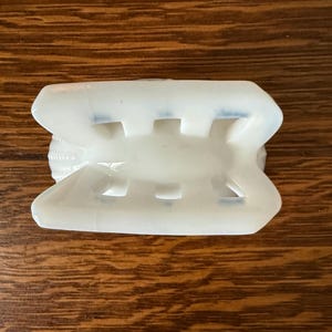 May include: A white, rectangular object with a series of rectangular cutouts. The object has a curved top and bottom, and appears to be made of a translucent material. The object is resting on a wooden surface.