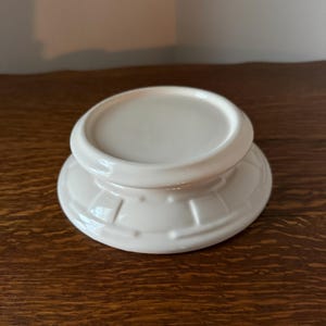May include: A white ceramic pedestal with a round top and a decorative, ridged base. The pedestal is a solid, off-white color and sits on a dark wooden surface. The design is simple and elegant.