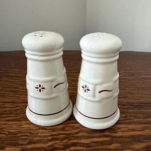 May include: A pair of off-white ceramic salt and pepper shakers with a rounded top and a tapered body. Each shaker features a dark red floral motif and a thin horizontal line near the base, with a series of small holes on the top for dispensing.