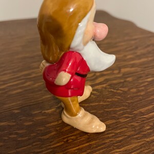 Może przedstawiać: Ceramiczna figurka krasnoludka z filmu Disneya Śnieżka i siedmiu krasnoludk&oacute;w. Krasnoludek ma na sobie czerwoną koszulę, brązowe spodnie i brązowe buty. Ma białą brodę i r&oacute;żowy nos.