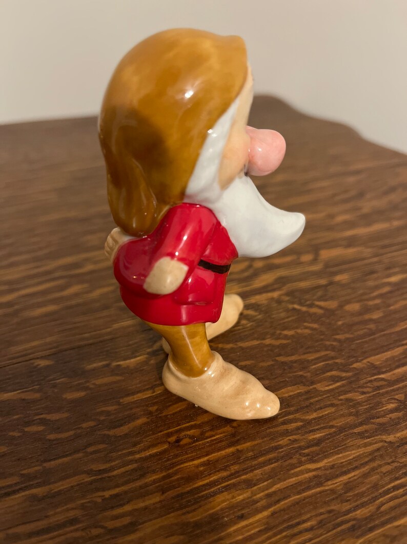 Może przedstawiać: Ceramiczna figurka krasnoludka z filmu Disneya Śnieżka i siedmiu krasnoludk&oacute;w. Krasnoludek ma na sobie czerwoną koszulę, brązowe spodnie i brązowe buty. Ma białą brodę i r&oacute;żowy nos.