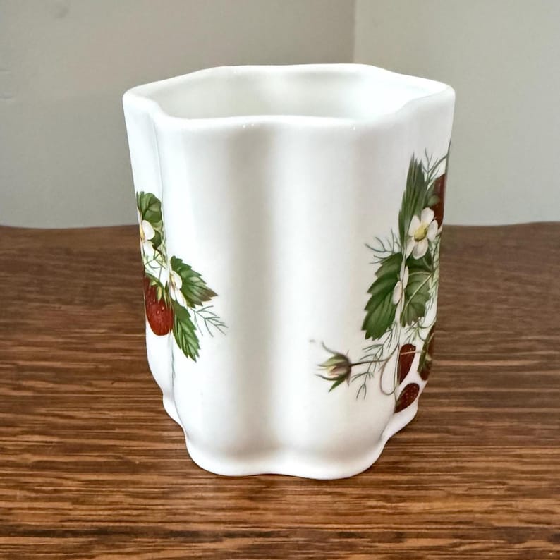 May include: A white ceramic tumbler with a scalloped, wavy rim and vertical fluted sides. The exterior features a botanical print of red strawberries, green leaves, and small white flowers arranged in clusters around the cylindrical body.
