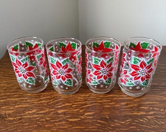 May include: Four clear drinking glasses with a festive poinsettia design. Each glass features red poinsettia flowers with green leaves and white snowflake accents. The glasses are arranged on a wooden surface.