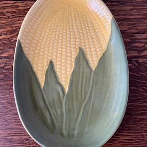 May include: A ceramic serving dish shaped like an ear of corn. The dish is green with yellow corn kernels and green leaves.