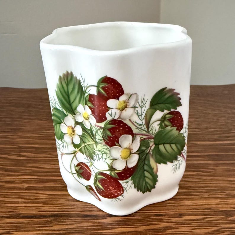 May include: A white ceramic tumbler with a scalloped rim featuring a botanical illustration of red strawberries, green leaves, and white flowers with yellow centers. The cylindrical vessel has a smooth, glossy finish and a slightly wavy top edge.