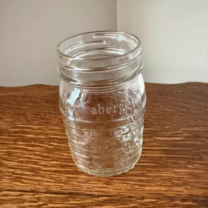 May include: A clear glass jar with a wide mouth and ribbed sides. The jar has the word "Waberge" embossed on the side. The jar is sitting on a wooden surface.