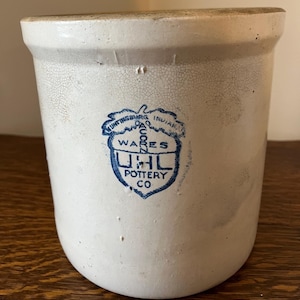 May include: A vintage off-white stoneware crock with a blue emblem that reads "Huntingburg, Indiana, UHL Pottery Co, Wares". The crock has a cylindrical shape with a slightly flared rim and a crackled surface.