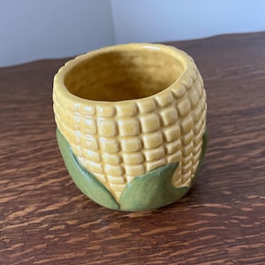 May include: A ceramic corn on the cob shaped planter with green leaves. The planter is yellow and has a textured surface.