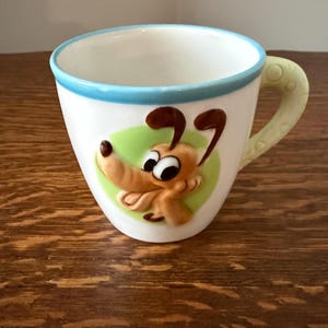 May include: A white ceramic mug featuring a raised, three-dimensional cartoon dog character on the front. The dog is light brown with dark brown ears and large eyes, set against a light green oval. The mug has a light blue rim and a pale green handle.